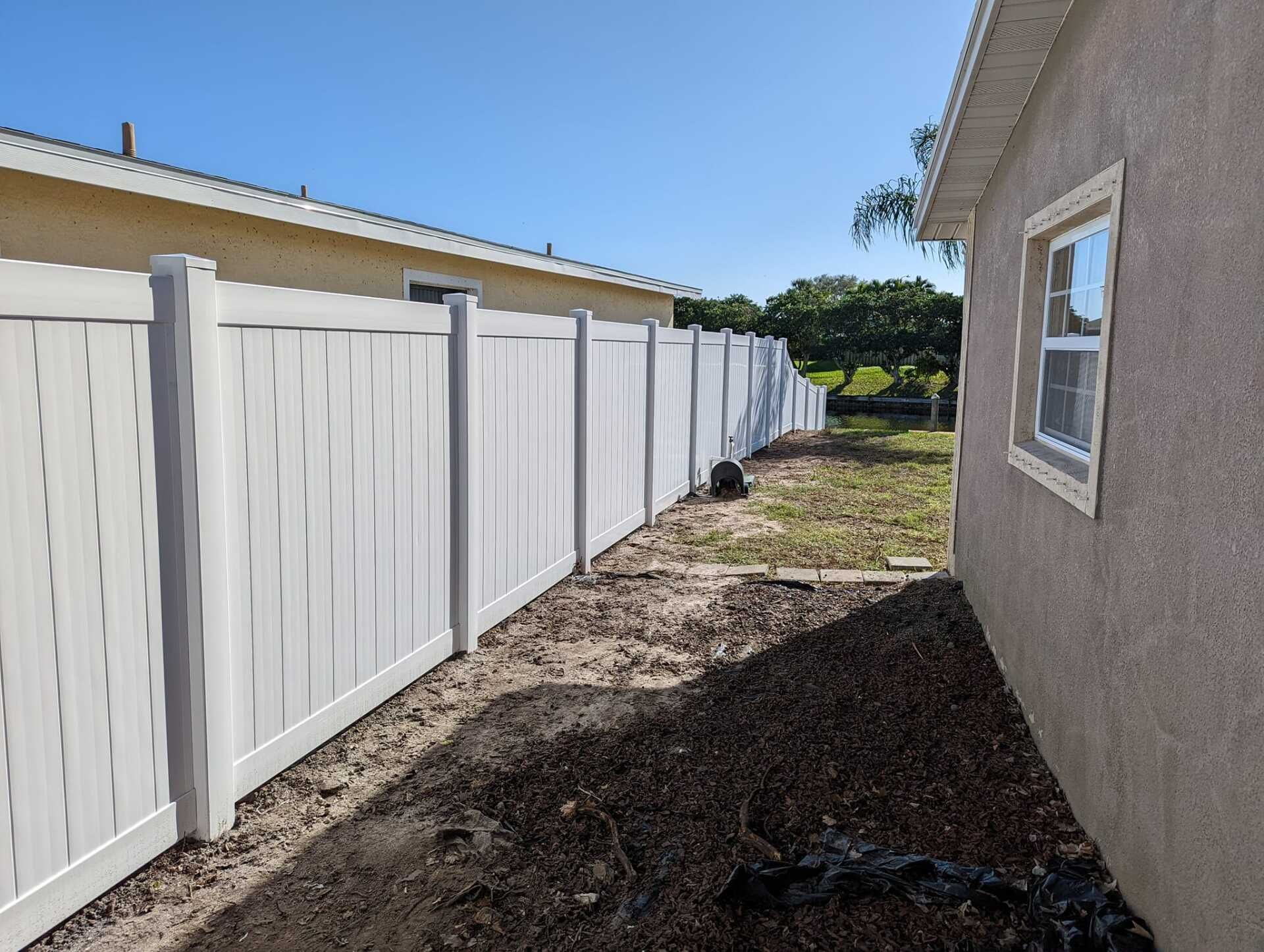 White vinyl fence along a tan house and bare ground, under a blue sky.