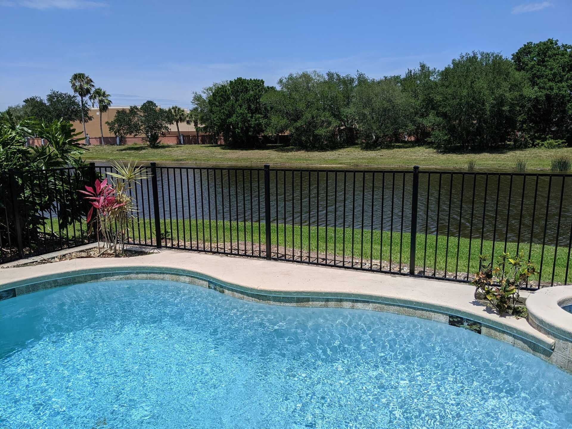 A swimming pool with blue water and a black fence overlooking a lake under a sunny sky.