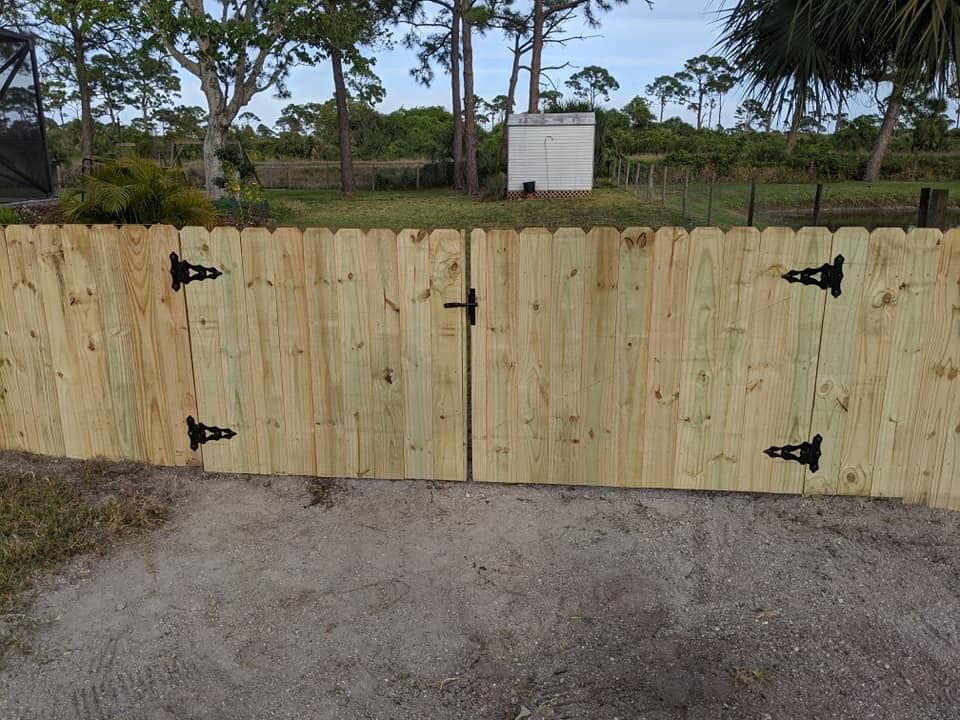 Wooden gate with black hinges and handle, set in a wooden fence, dirt driveway.