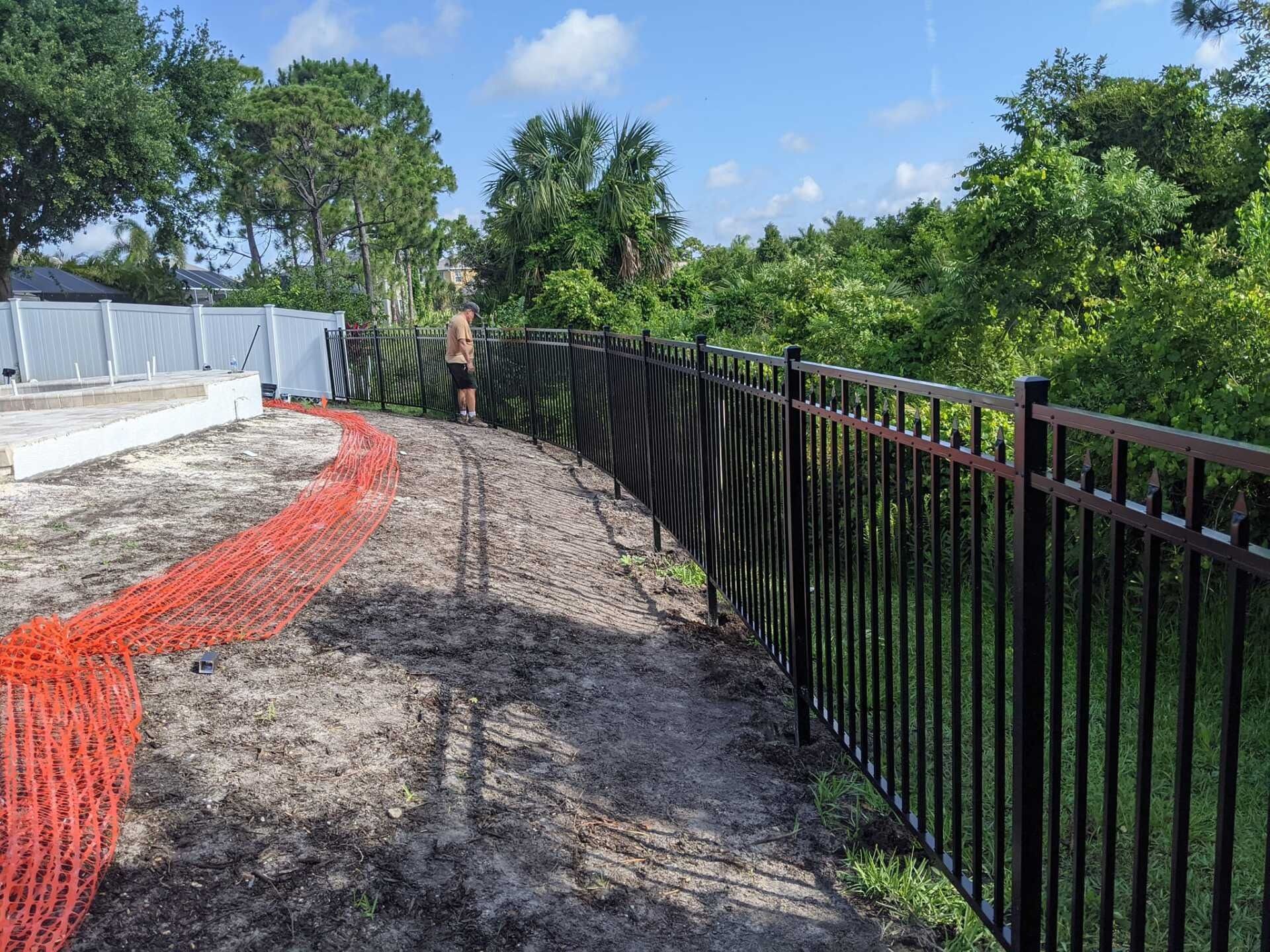 Black metal fence along a dirt path with orange construction fencing. A person stands nearby. Trees and blue sky in the background.