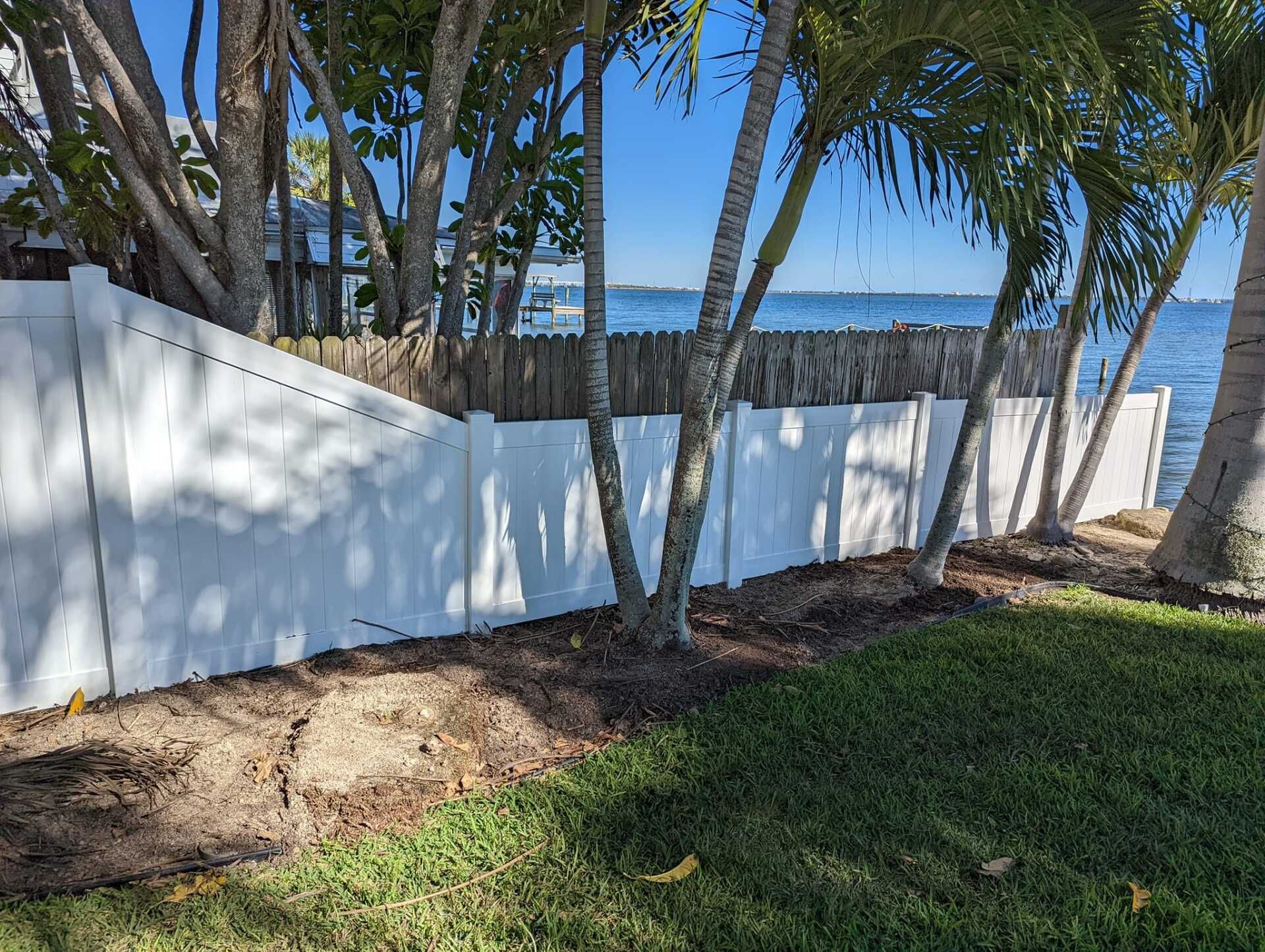White fence along the water's edge with palm trees and a view of the sea under a blue sky.