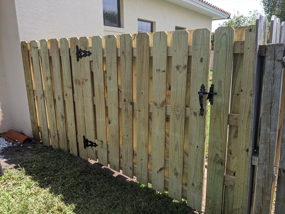 Wooden picket fence with gate, set next to a beige building, with green grass in front.