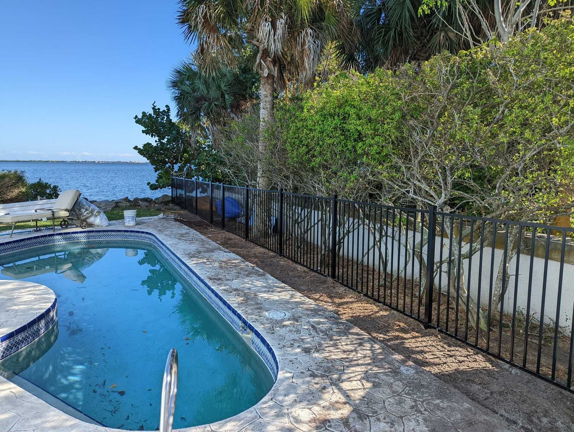 Poolside view: a blue pool with clear water, black fence, and trees near a body of water under a blue sky.