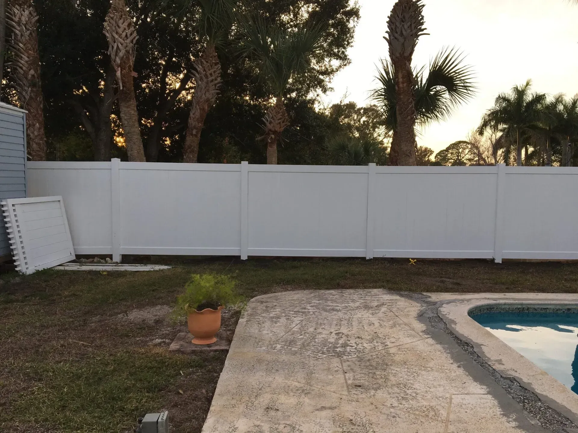 White privacy fence along a backyard with a pool. Palm trees and a cloudy sky in the background.