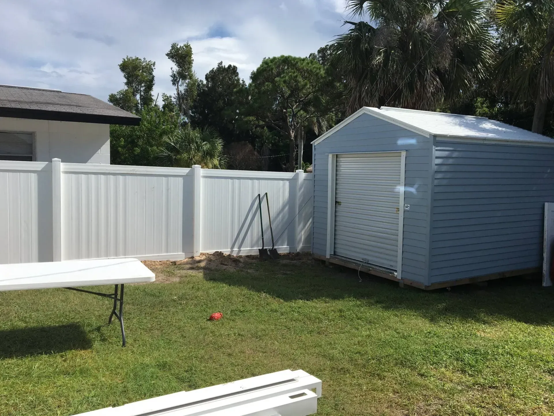 Backyard scene: white fence, blue shed with garage door, white table, shovels, green grass.