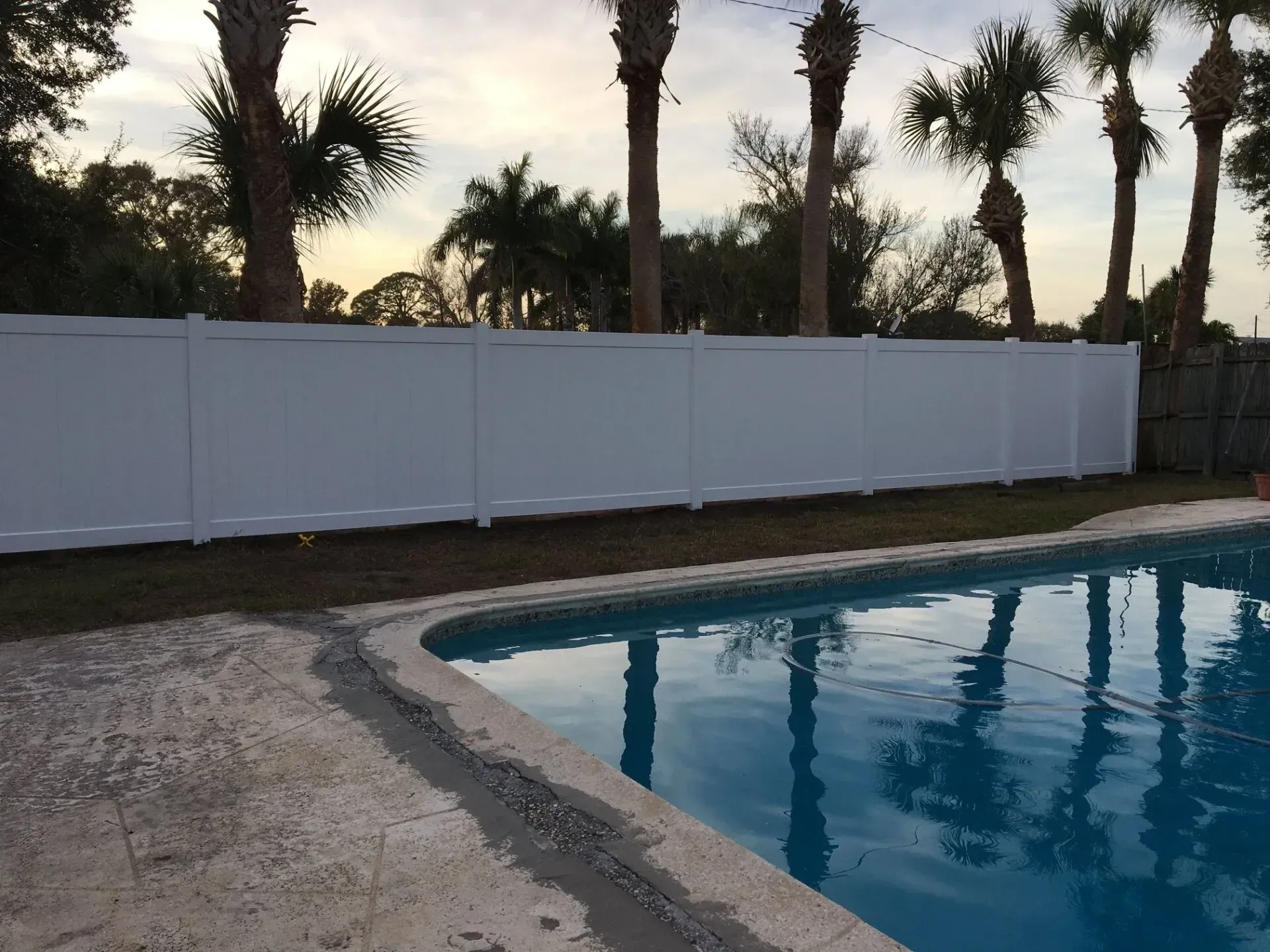 White fence bordering a swimming pool, with palm trees in the background under a dusky sky.