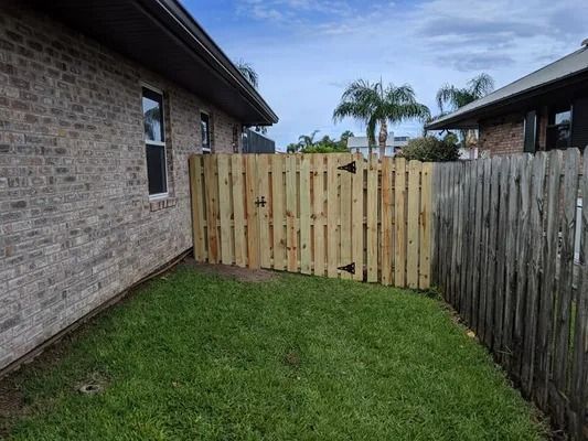 Wooden fence separating brick building and weathered fence in a grassy yard under a cloudy sky.