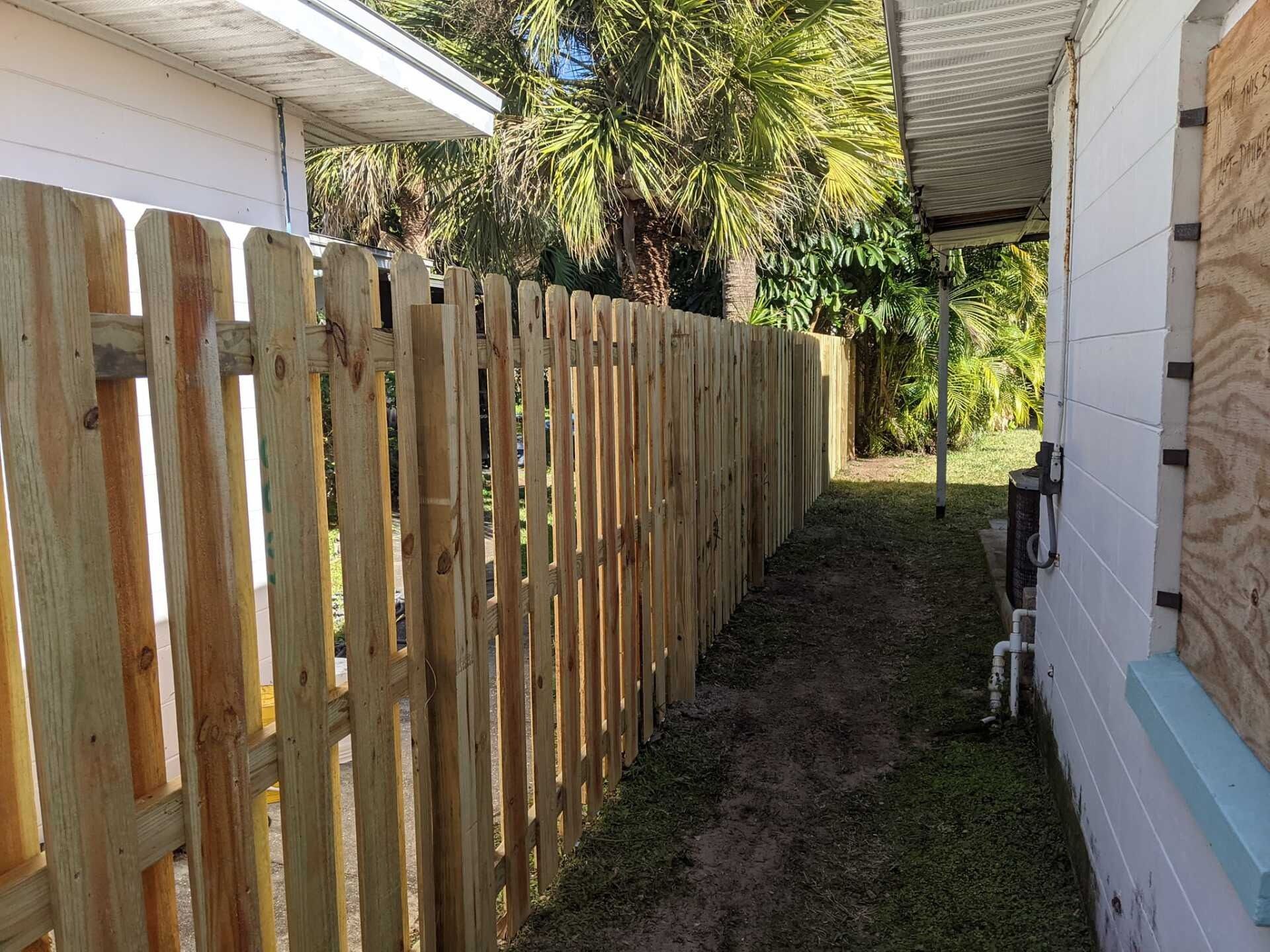 Wooden picket fence next to a white house with overgrown grass and lush green trees.
