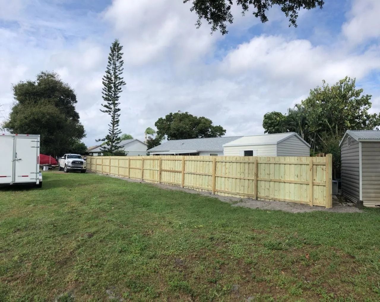 A light-colored wooden fence runs along the edge of a grassy yard with houses in the background under a cloudy sky.
