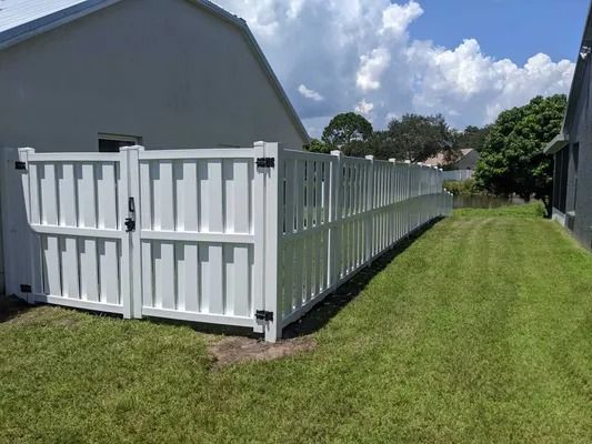 White vinyl fence with gate, bordering a grassy yard, next to a house under a blue sky.