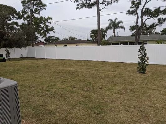 White fence encloses a grassy backyard with trees under an overcast sky.