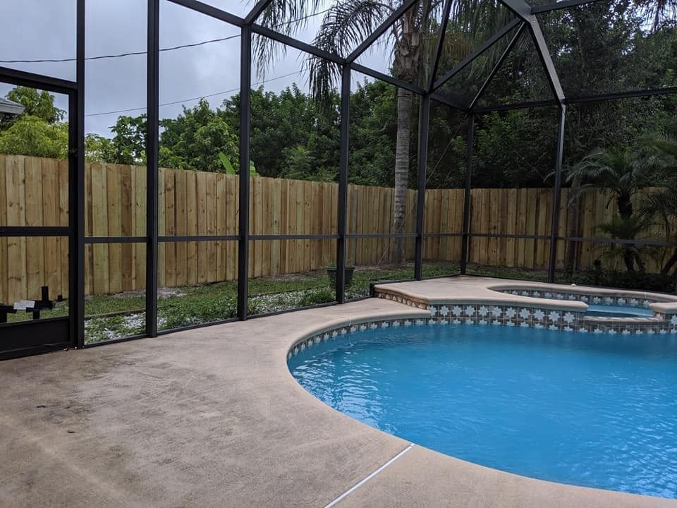 Pool area with screen enclosure and wooden fence. Blue pool, concrete deck, and green trees.