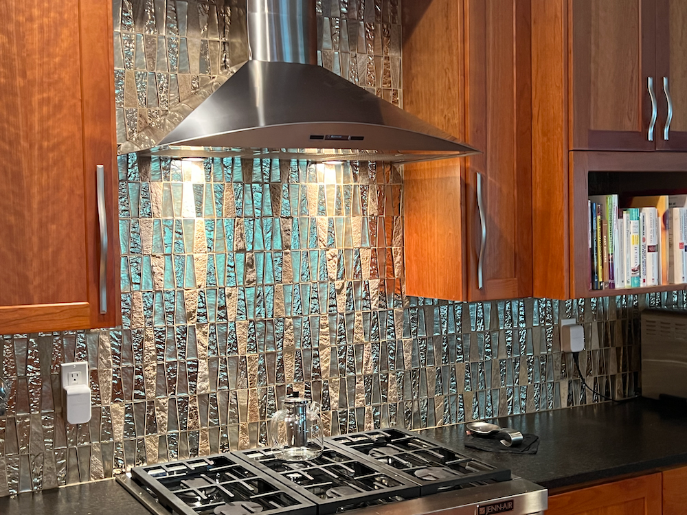 Kitchen backsplash with mirrored tiles and stainless steel range hood above a gas cooktop.