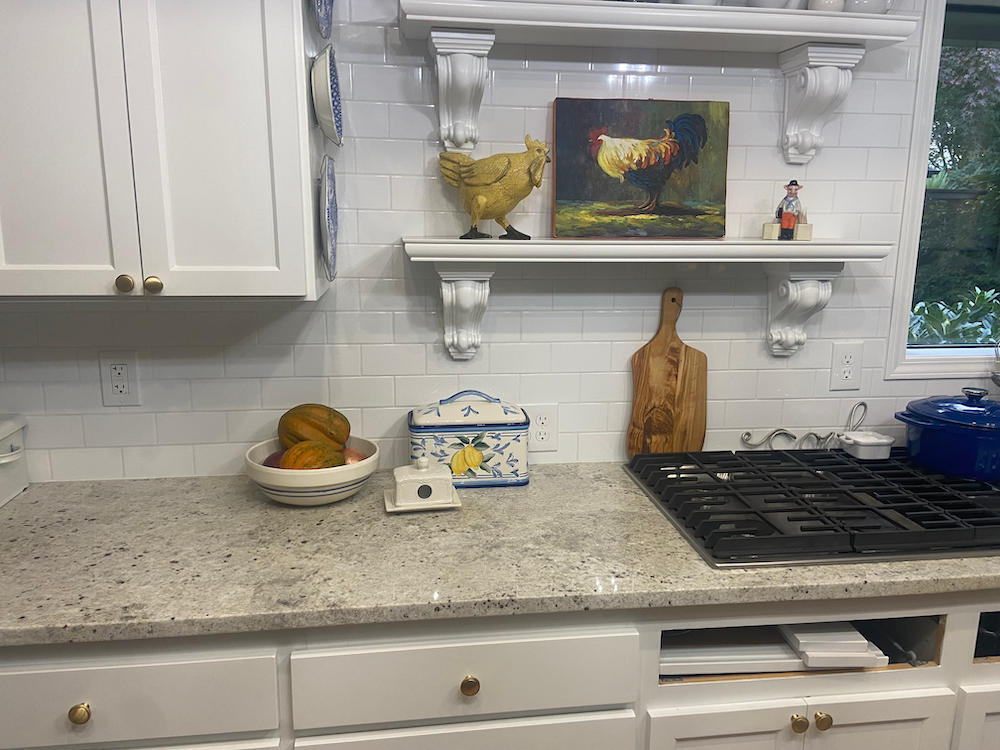 White kitchen with granite countertops, decorated with chicken art and a wooden cutting board.