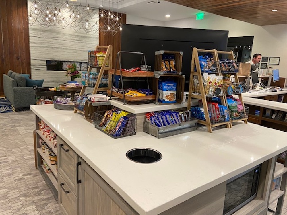Snack display on a counter. Chips, cookies, and other items for sale. Wooden shelves, neutral tones.