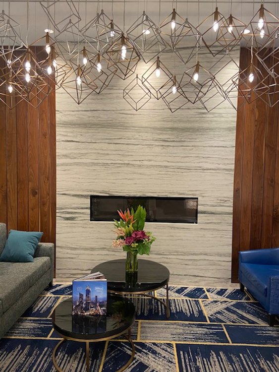 Lounge area with fireplace and geometric lighting fixtures. Gray and blue seating, coffee tables, and floral arrangement.