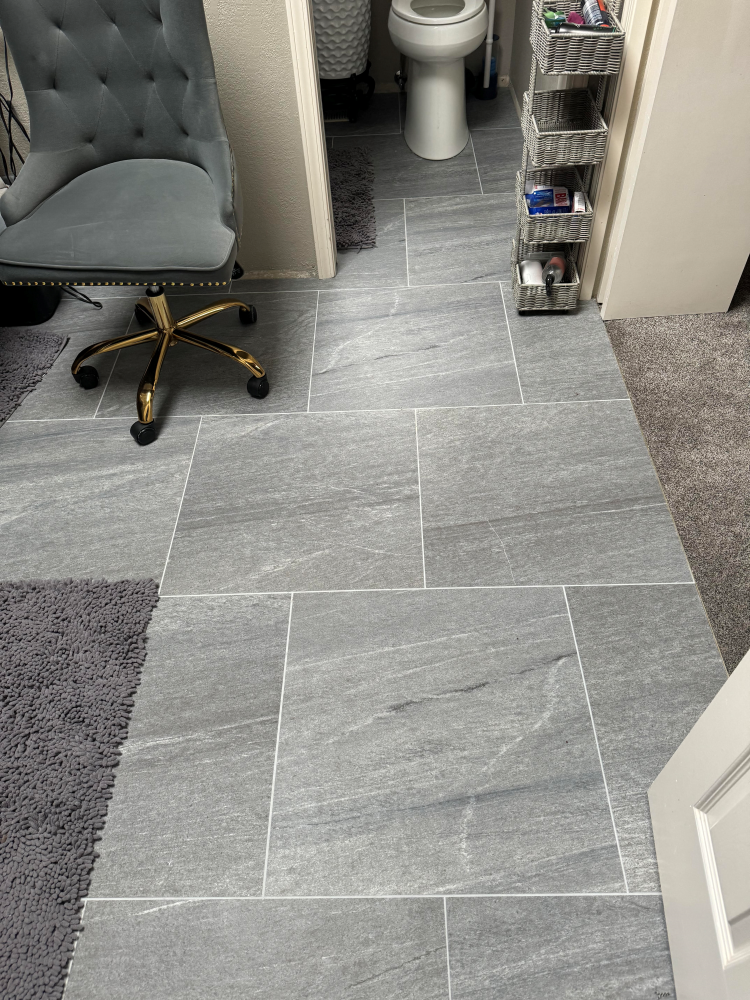 Gray tiled bathroom floor with a chair, rug, and doorway to the toilet.