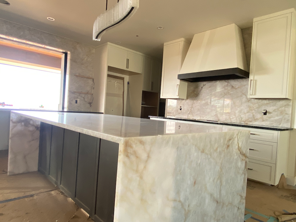 Modern kitchen with large island and marble countertops. Gray cabinets, white range hood, and neutral wall tile.