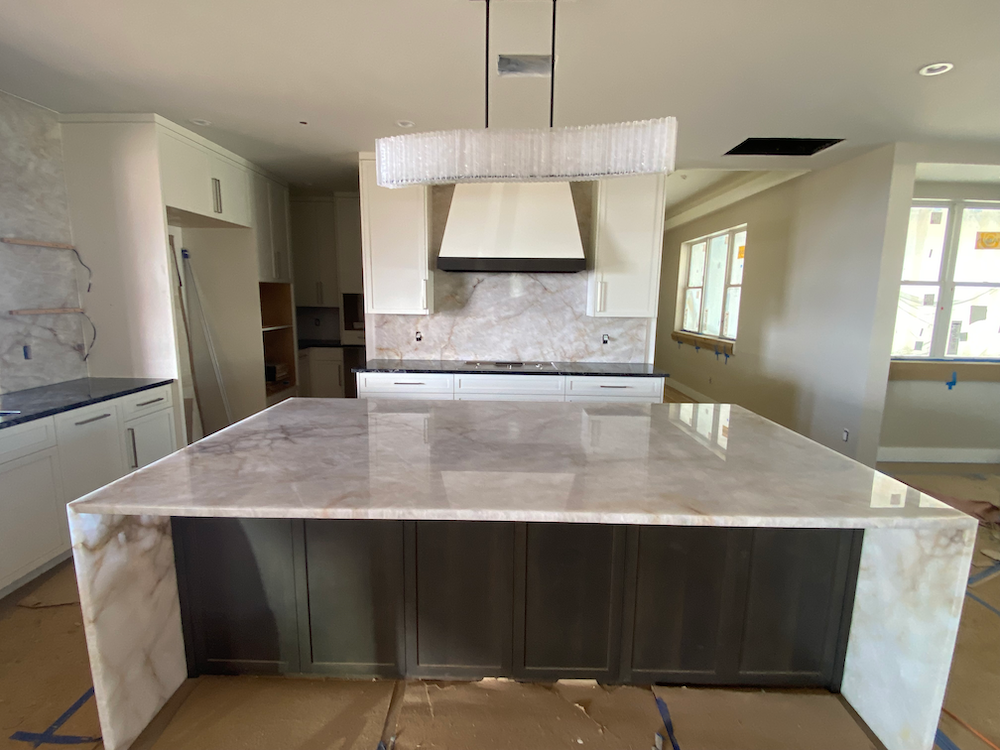 Modern kitchen with large island, marble countertops, dark cabinets, and light fixture.