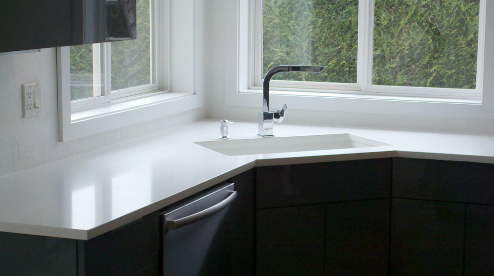 Kitchen corner with white countertop, dark cabinets, stainless steel faucet, and window overlooking trees.