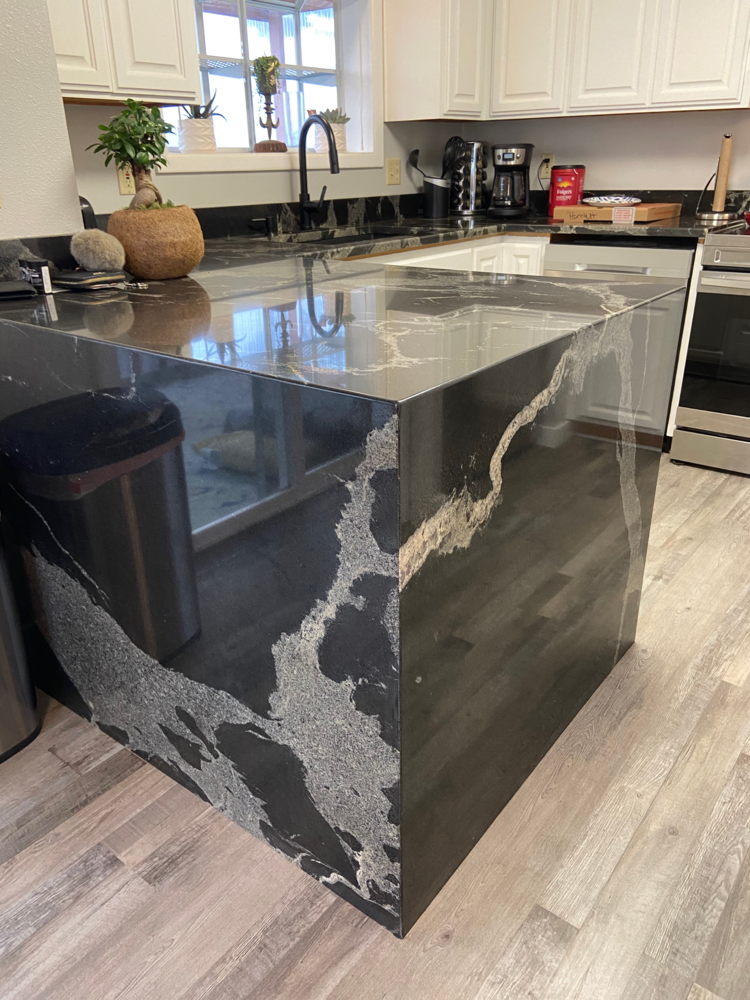 Kitchen island with black and white veined countertop, stainless steel trash can, and light wood flooring.