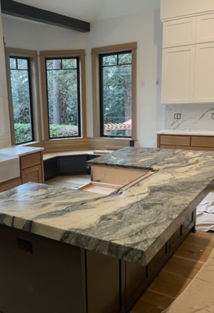 Kitchen with large marble island, wood cabinets, and bay window looking out onto trees.