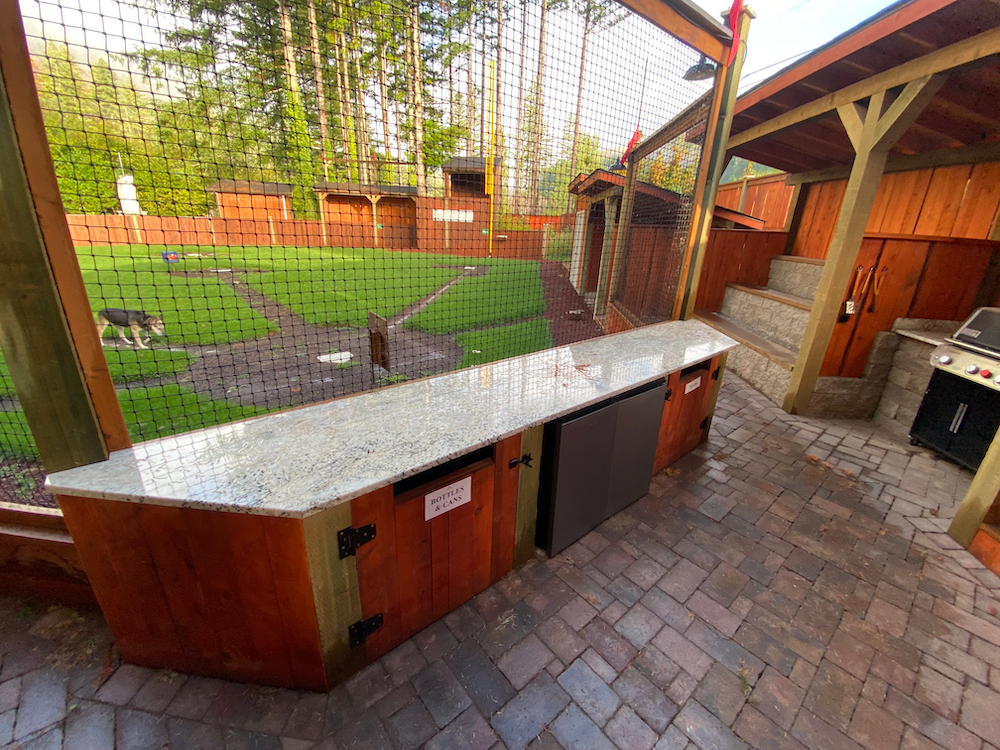 Outdoor bar with granite countertop, built-in fridge, and wooden cabinets.  Brick patio leads to fenced yard with dog.