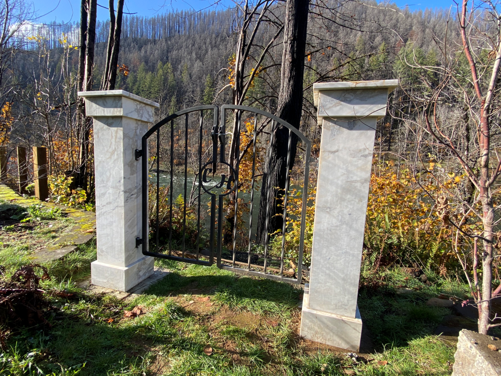Wrought iron gate between two stone pillars, set in a grassy area with trees and a view of a forested hillside.