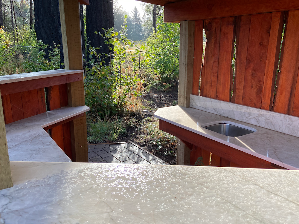 Outdoor kitchen structure with countertops, sink, and view of trees.