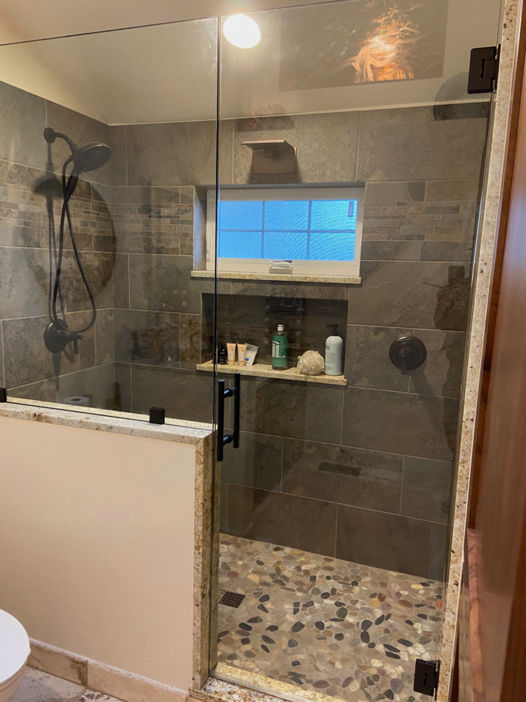 Modern shower with glass door, stone tile walls and pebble floor. Includes shelf with toiletries and window.