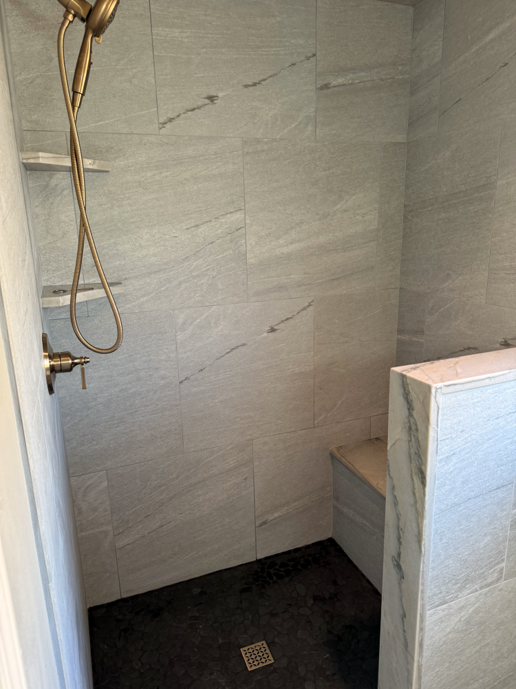 Shower with gray tiled walls, black floor, golden fixtures, and a built-in bench.