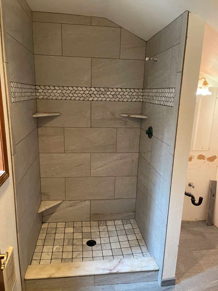 Newly tiled shower with gray walls, mosaic accent, and marble floor, showcasing shelves and fixtures.