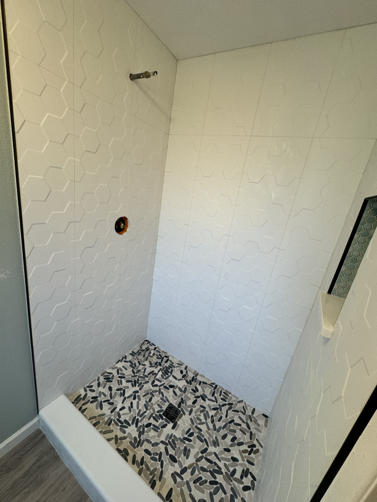Small shower with white honeycomb-patterned tiles, stone pebble floor, and glass door.