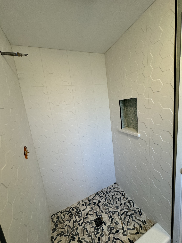 White tiled shower stall with hexagon pattern on walls, mosaic floor, and built-in shelf.