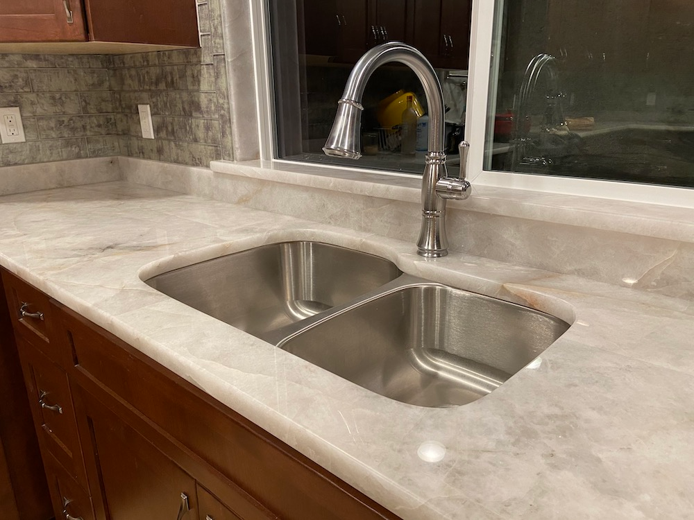 Stainless steel double sink and faucet in kitchen, with light-colored countertops and dark wood cabinets.