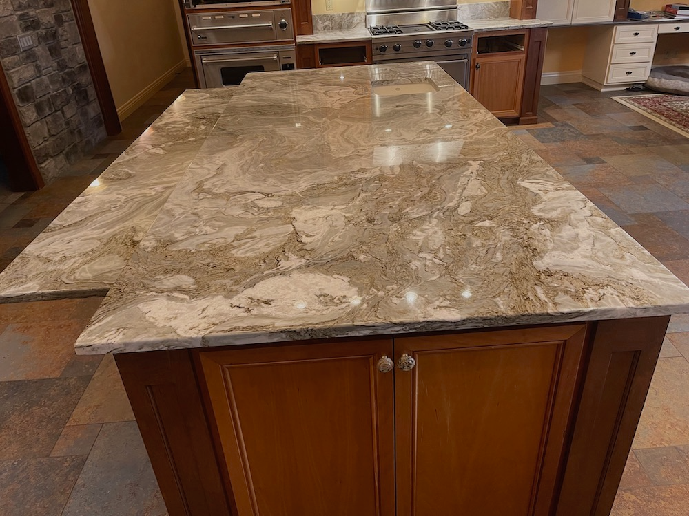 Granite countertop on a brown cabinet island in a kitchen, stainless steel appliances visible.