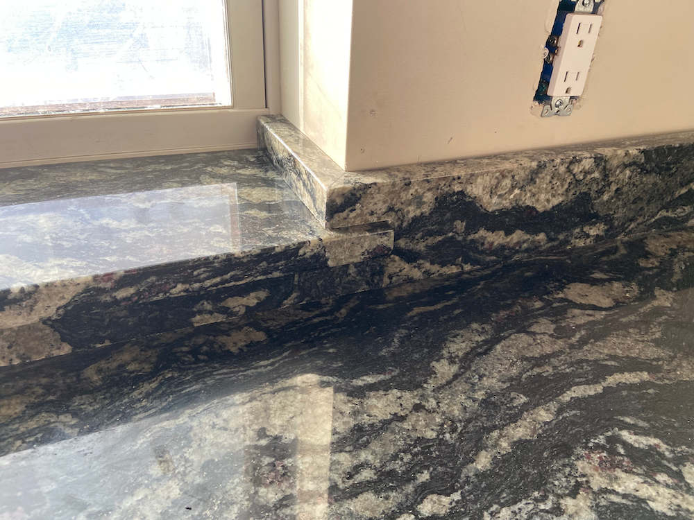 Granite countertop with dark veining meets a window sill and a wall with an electrical outlet.