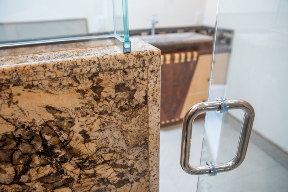 Granite countertop with glass shower door and stainless steel handle. Beige and brown tones.