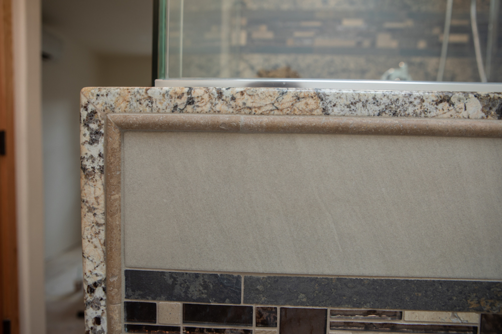 Close-up of a bathroom counter with granite edging and textured tile with a mosaic border.