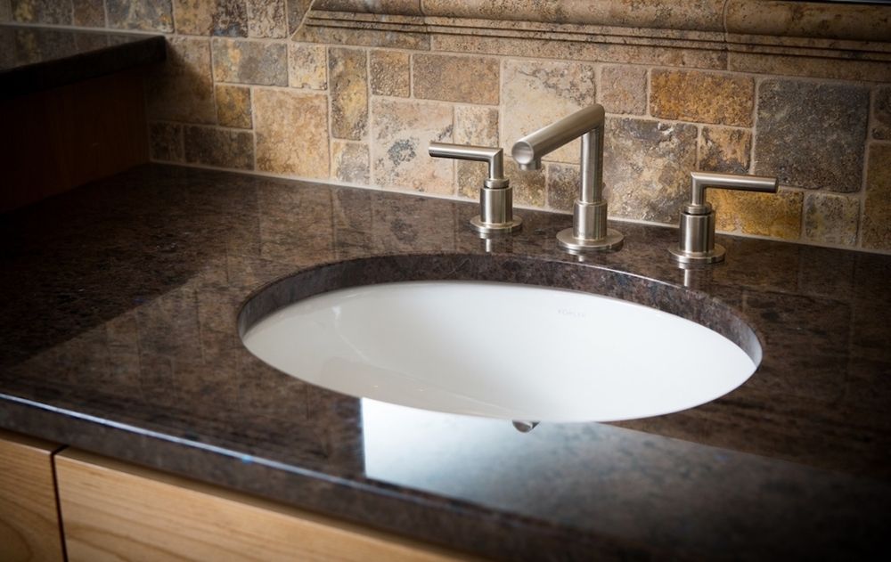 Oval white sink in a dark brown countertop with a silver faucet and tile backsplash.