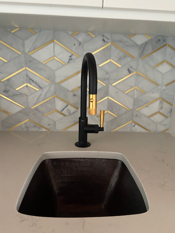 Copper sink with black and gold faucet against a marble and gold tile backsplash.