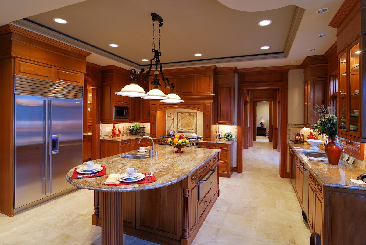 Luxurious wooden kitchen with granite countertops, island with sink and pendant lights, stainless steel refrigerator.