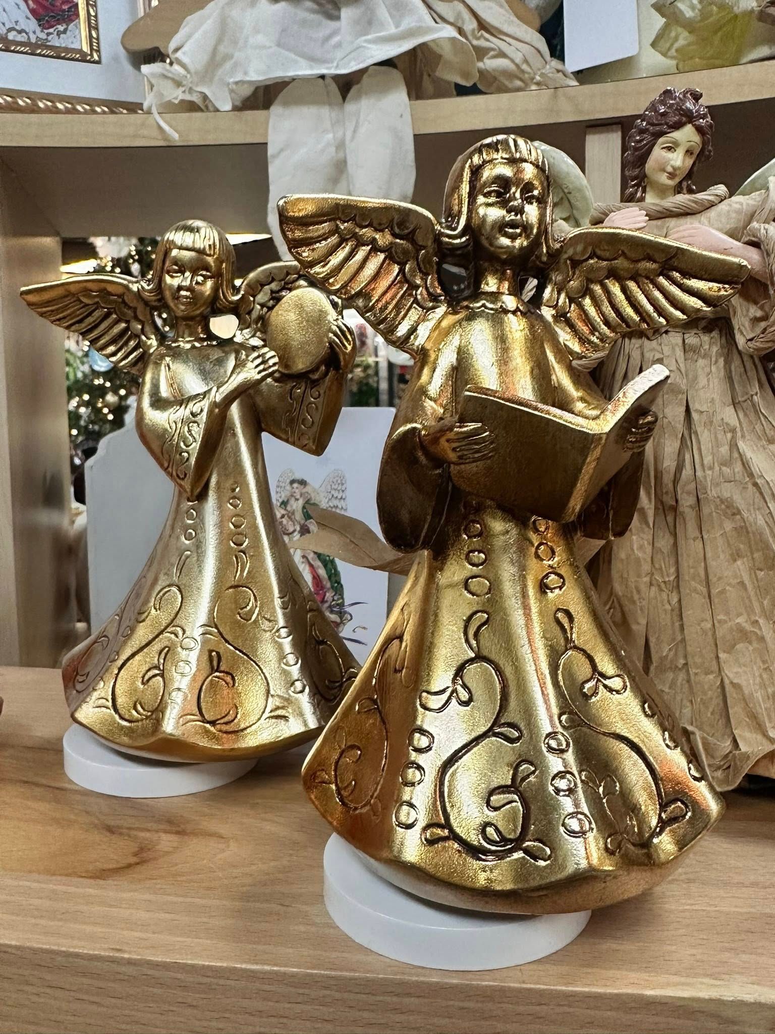 Two golden angel figurines with raised wings, holding a book and ball, on a wooden shelf.