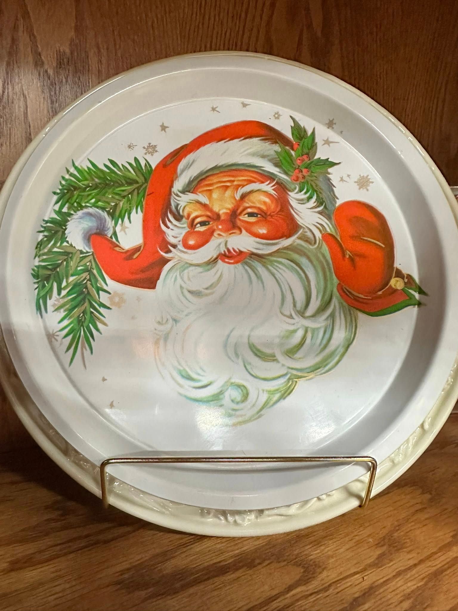 Round white metal serving tray with a Santa Claus illustration; gold-colored wire stand.