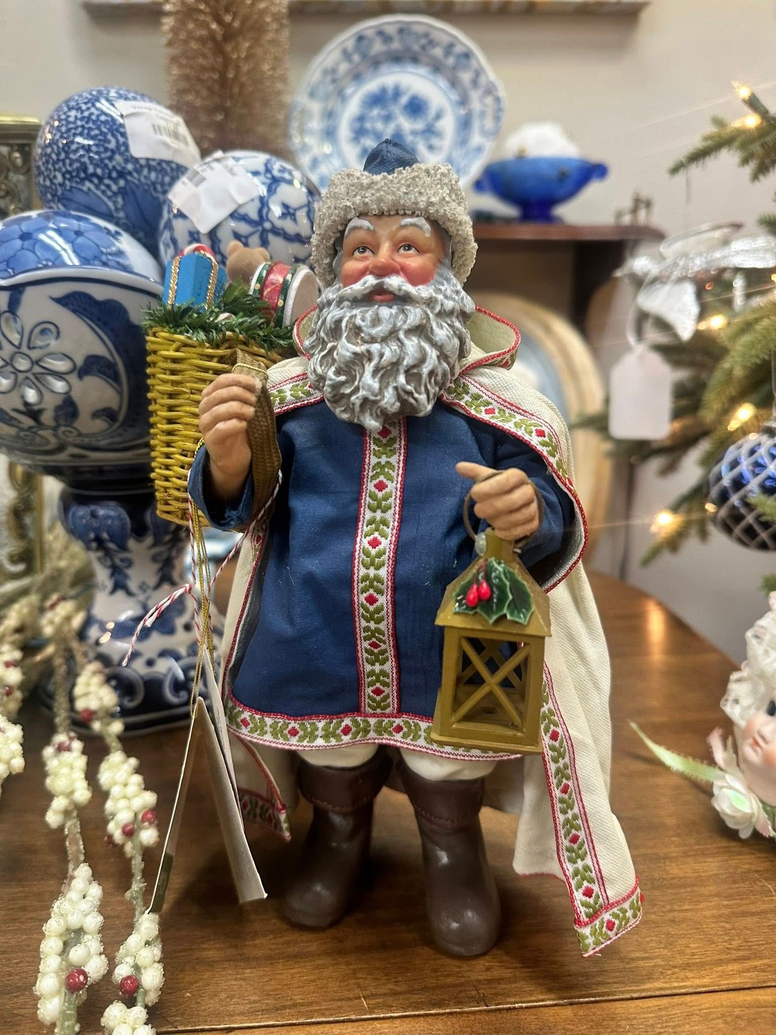 Santa figurine holding a lantern and basket, wearing blue coat with white trim, brown boots, setting with other decorations.