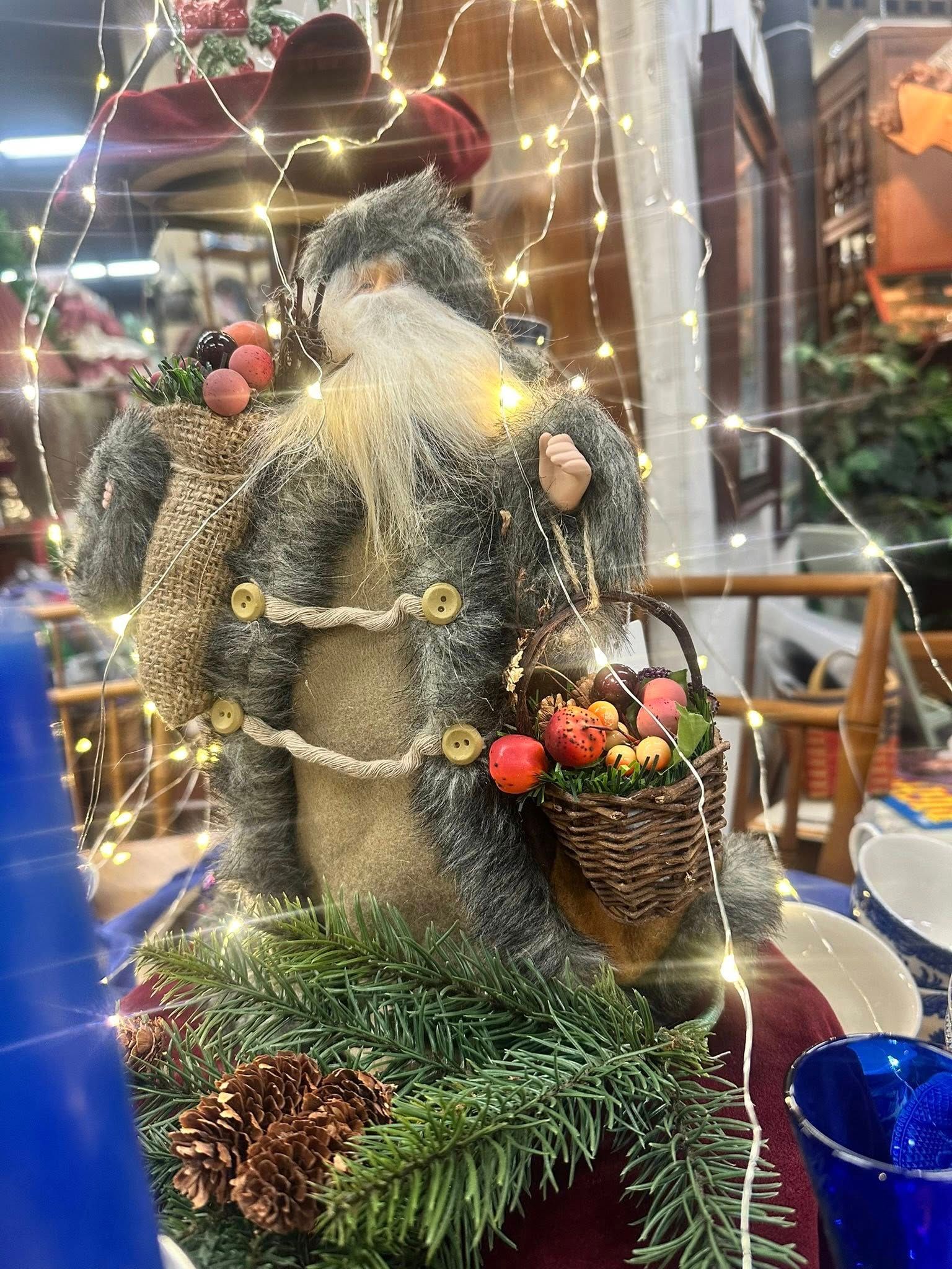 Santa Claus figurine with string lights, carrying baskets of flowers, set on greenery.