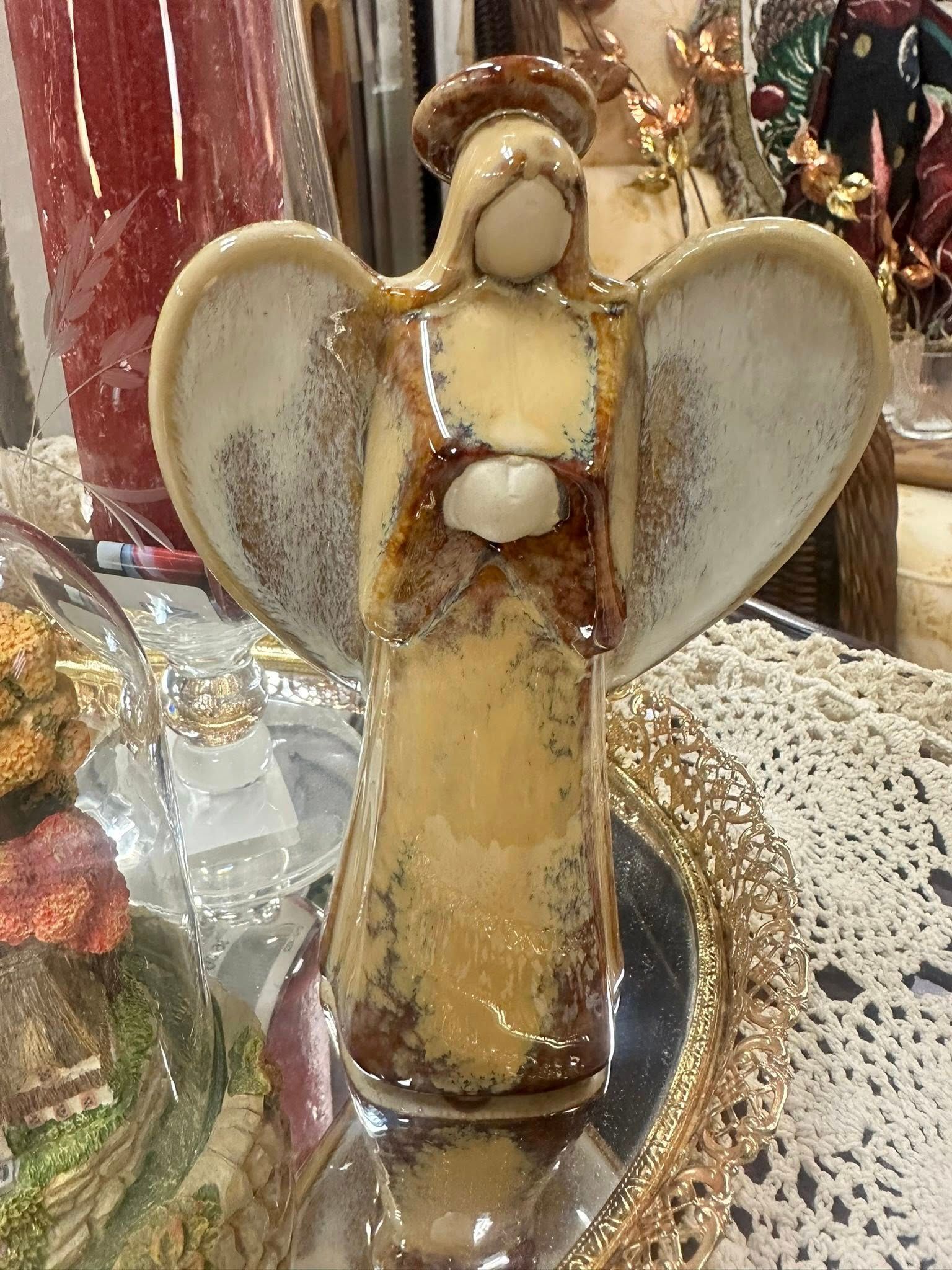 Ceramic angel statue with tan wings and dress, halo, holding a small object, standing on a mirrored surface.