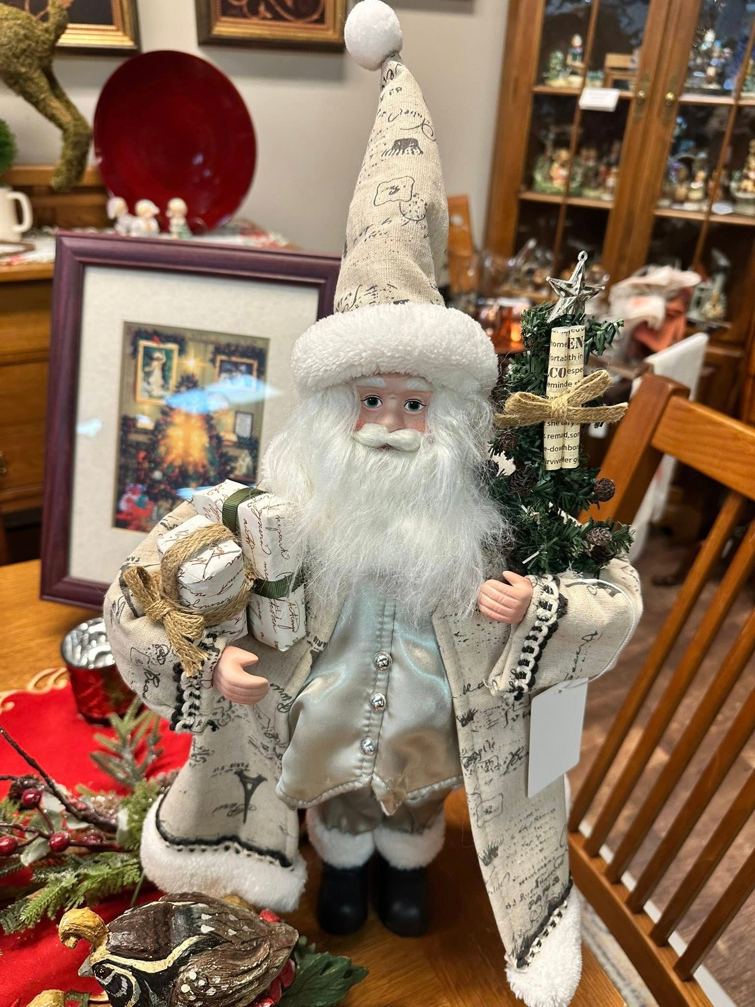 Santa Claus figurine holding presents and a tree, dressed in a winter coat, standing indoors.