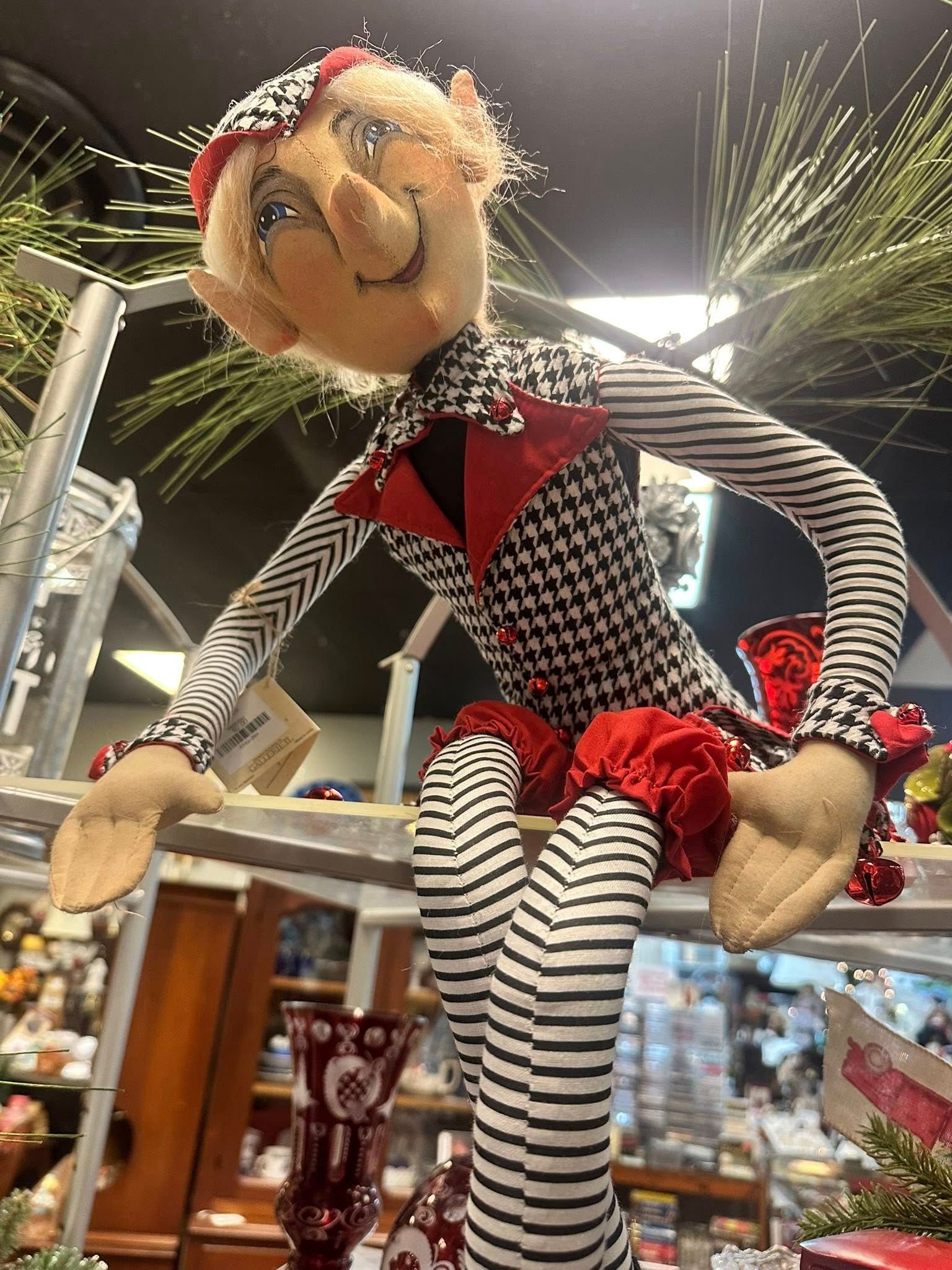 Elf doll in houndstooth jacket and striped legs, sitting on a metal bar, decorative Christmas setting.
