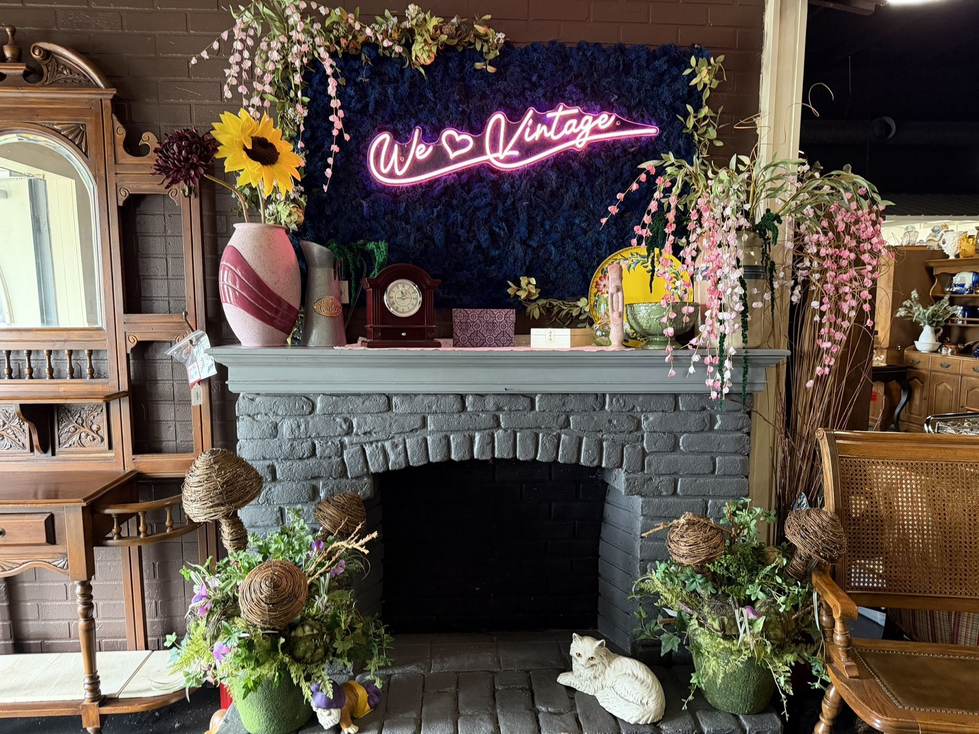 Cozy rustic mantel with neon sign, flowers, and pumpkins beneath a gray brick fireplace